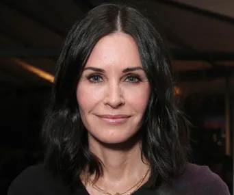 Courteney Cox smiling with black hair, wearing a necklace, indoor setting.
