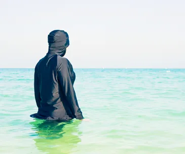 Person wearing a black burkini stands in the sea, facing the horizon.