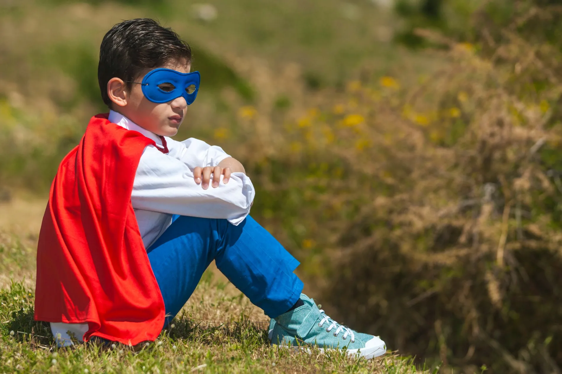 Child in superhero costume with blue mask and red cape sitting on grass, arms crossed, looking thoughtful.