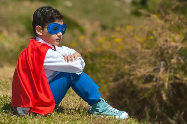 Child in superhero costume with blue mask and red cape sitting on grass, arms crossed, looking thoughtful.