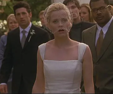 Bride in veil looks surprised, standing with a wedding group, including people in suits and dresses in an outdoor setting.