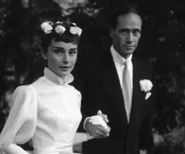 A black-and-white photo of a bride in a white dress with floral headpiece, holding hands with a man in a suit with a boutonniere.