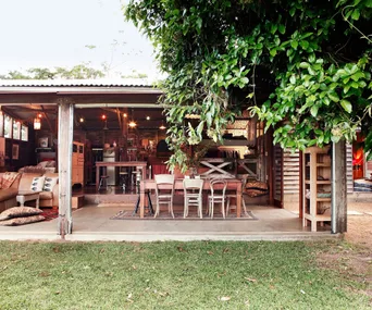Rustic open garden room with wooden furniture, plants, a cozy couch, dining table, and green lawn outside.