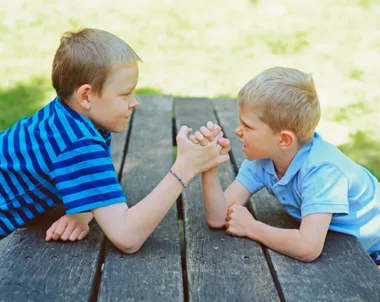 Two young boys arm wrestling on a wooden table outdoors, both focused and determined.