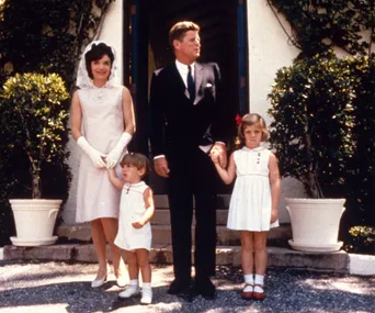 A family poses in front of a building with two children in white dresses holding their parents' hands.