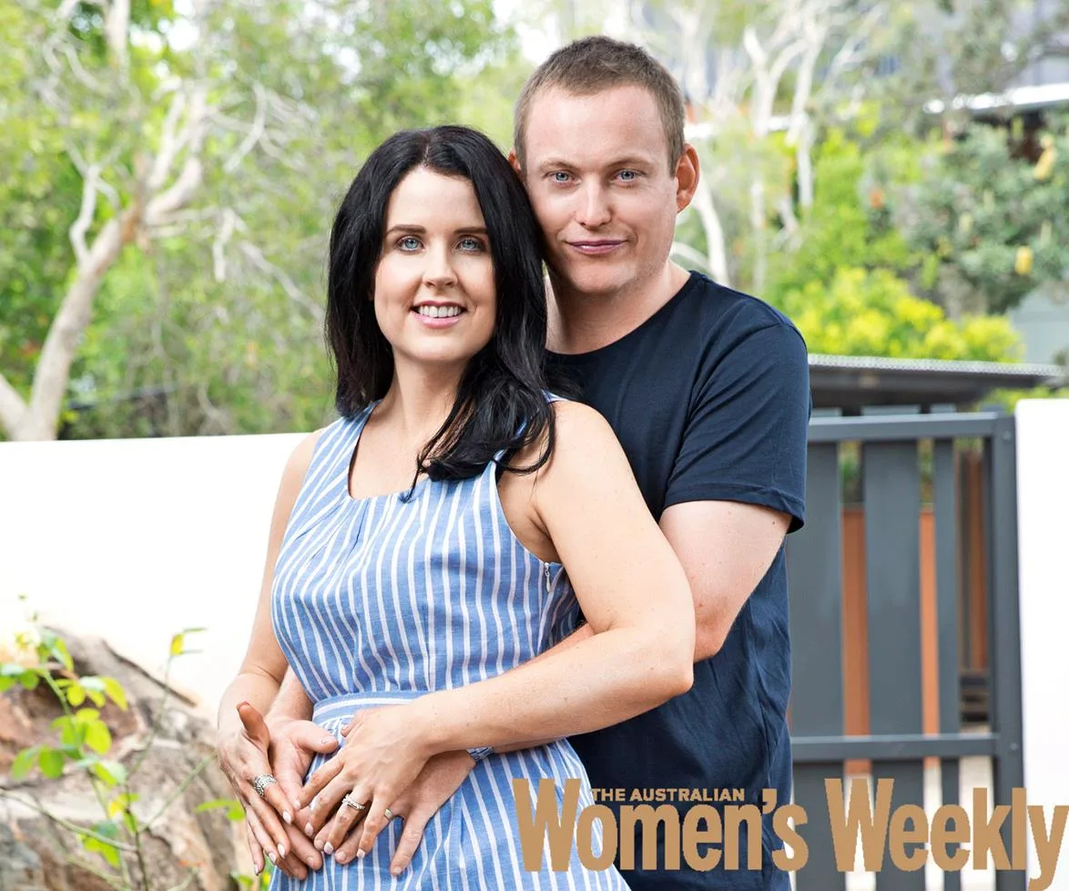 A couple smiling outdoors, with the woman in a striped dress and the man in a navy shirt, hands on her belly.