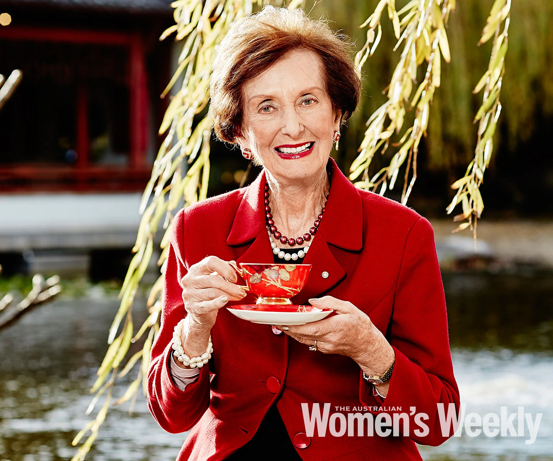 Elderly woman in red blazer holding a floral teacup outdoors by the water.