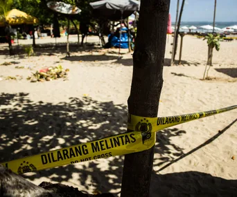 Yellow "Do Not Cross" tape on a sandy beach with trees and umbrellas in the background.