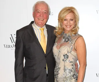A man in a suit and a woman in a floral gown smiling together on a red carpet.