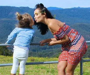 Mother and daughter playfully kissing outdoors on a sunny day with a scenic mountain background.