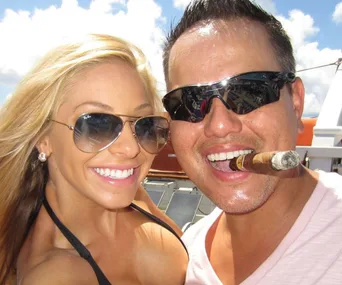 A smiling couple in sunglasses with the man holding a cigar in his mouth.