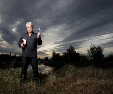 A man holding a Bible and a cross stands in a field under a dramatic cloudy sky.