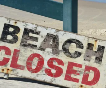 "Weathered 'Beach Closed' sign on a sandy shoreline with a metal railing in the background."