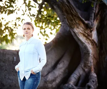 A woman in a white shirt and jeans stands by a large tree, with a soft background of leaves and light.
