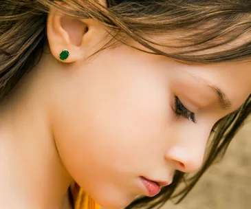 A young girl in profile wearing a green stud earring, with closed eyes and brown hair.