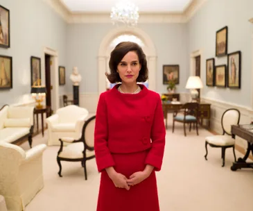 Natalie Portman as Jackie Kennedy in a red suit, standing in an elegantly furnished room.