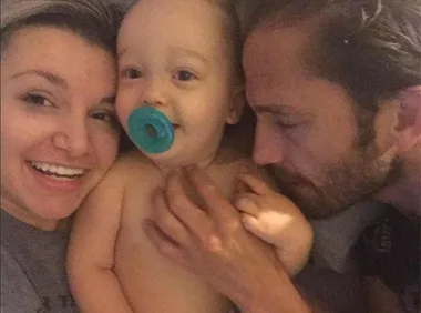 A family selfie with a smiling woman, a baby with a pacifier, and a man lying close, conveying warmth and togetherness.