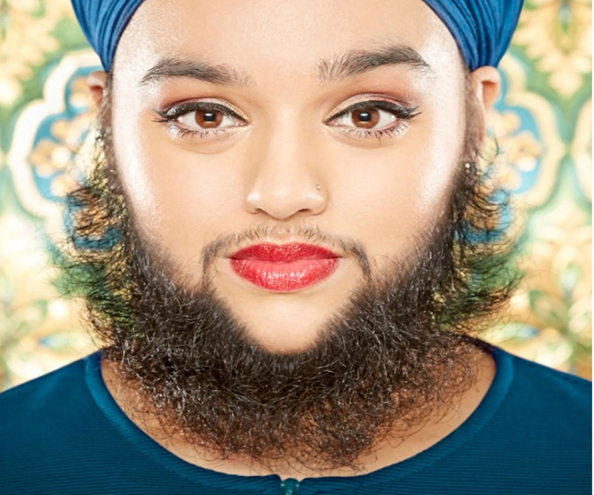 Woman wearing blue turban and red lipstick, with a beard, in front of a decorative background.