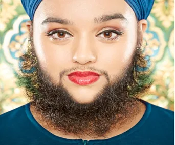 Woman wearing blue turban and red lipstick, with a beard, in front of a decorative background.