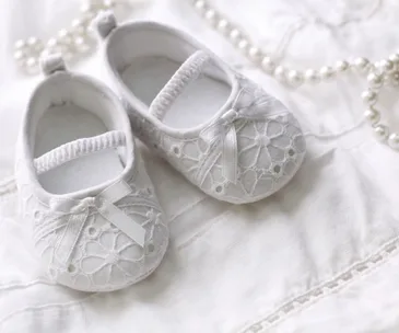 White baby shoes with lace detail and satin ribbon on a white fabric background with pearls.