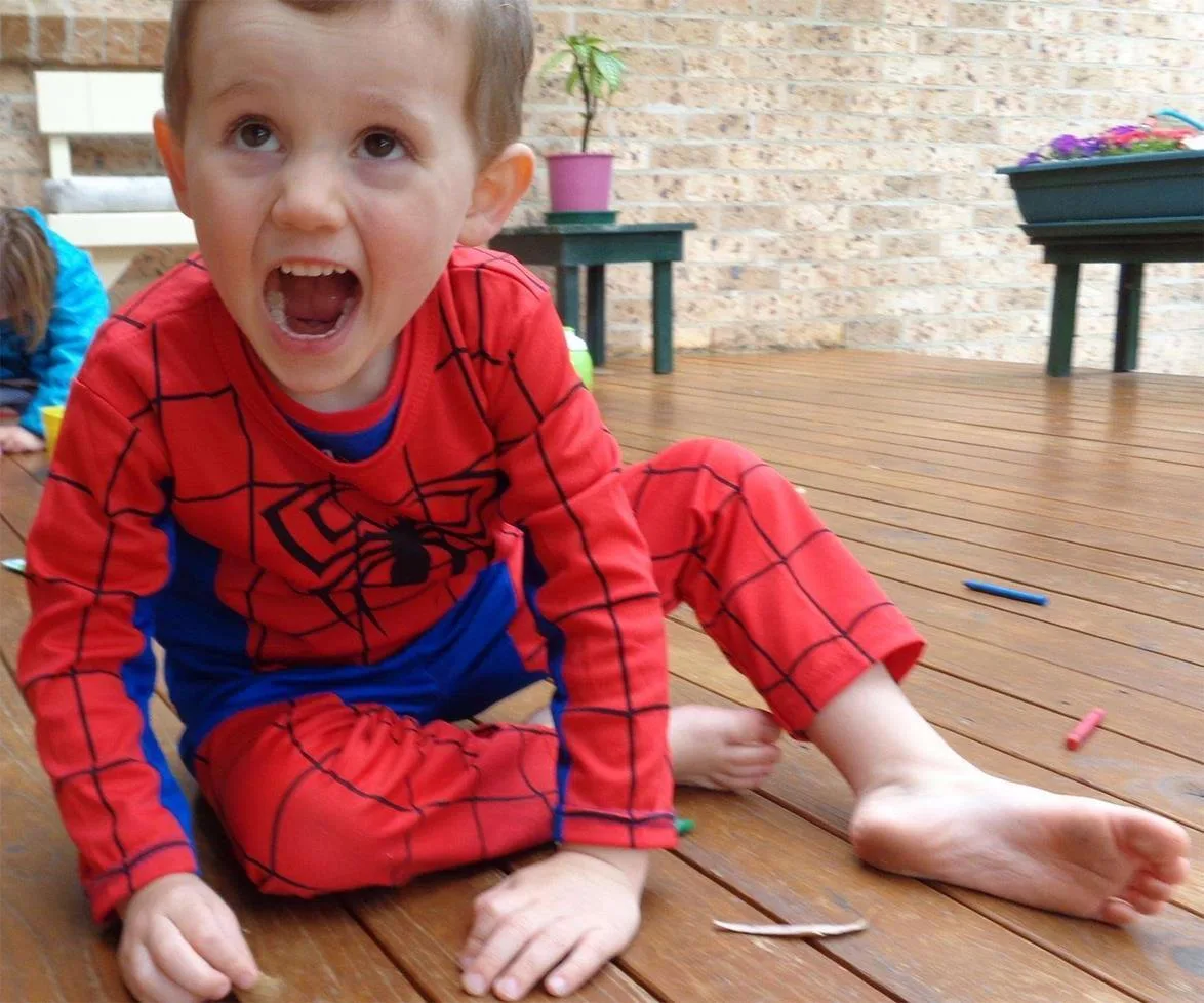 Young child in a Spider-Man costume sits on a wooden deck, smiling widely, with colorful chalk pieces scattered around.