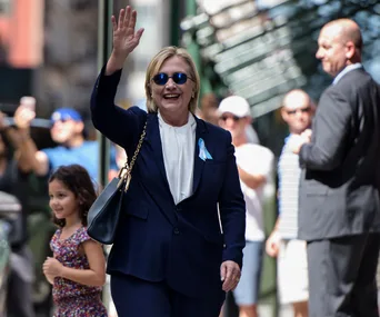 A woman in a blue suit waves while walking outside, surrounded by people.