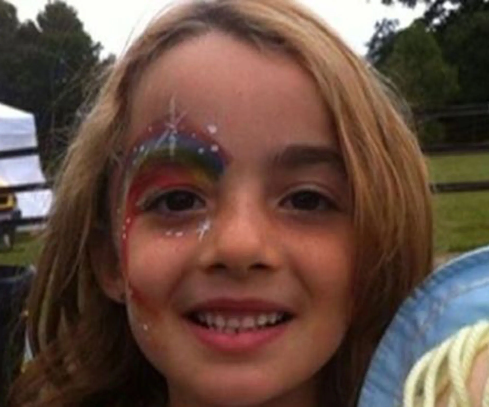 A young girl with face painting, smiling outdoors, rainbow design around her eye.