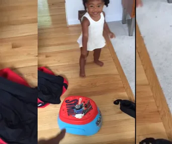 Young child in white dress smiling near a "Thomas the Tank Engine" potty training seat on a wooden floor.