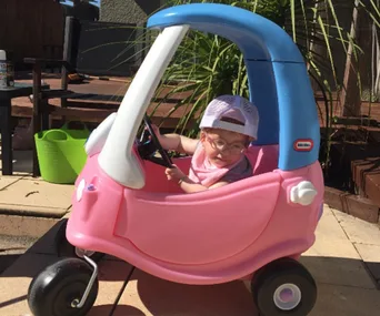 A child in a pink and blue toy car wearing a cap and glasses, outdoors near a wooden deck.