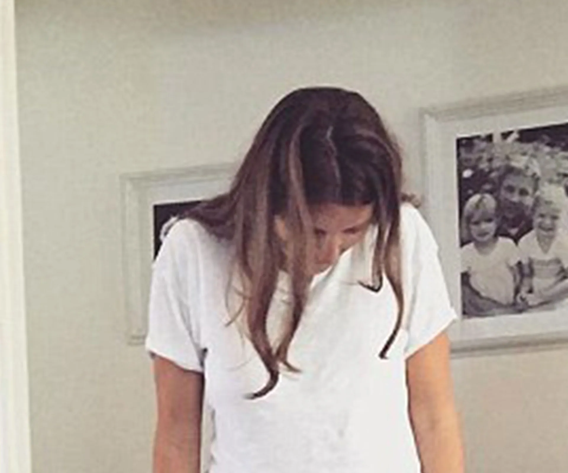 Woman in white T-shirt looking down, with framed family photos on wall behind her.