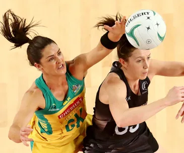 Players from Australia and New Zealand compete in a netball match, intensely reaching for the ball mid-air.