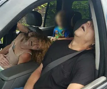 Adults appear unconscious in a car while a child sits in the backseat, blurred.