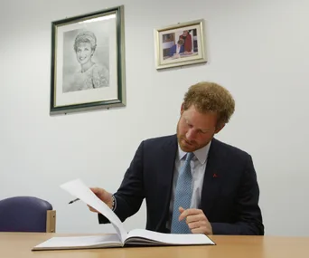 Prince visiting hospital, signing book under Diana's portrait, wearing suit with red ribbon pin.