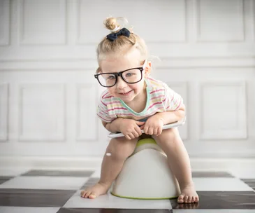 A toddler in glasses sits on a potty chair, smiling, with a top knot hairstyle and a colorful striped shirt.