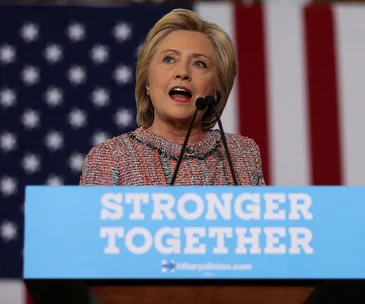 Hillary Clinton speaks at a podium with "Stronger Together" sign, U.S. flag backdrop.