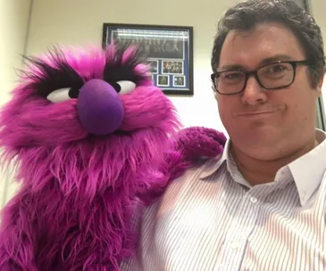 A person with glasses poses with a large purple and pink furry puppet in an office setting.