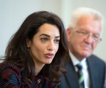 Amal Clooney speaking at a press event with a man in a suit sitting beside her in a blurred background.