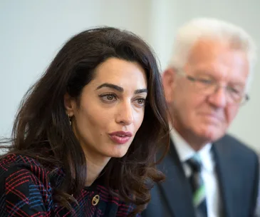 Amal Clooney speaking at a press event with a man in a suit sitting beside her in a blurred background.