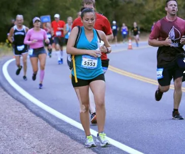 A woman, with bib number 5553, multi-tasks by pumping breast milk while running a half-marathon on a winding road.