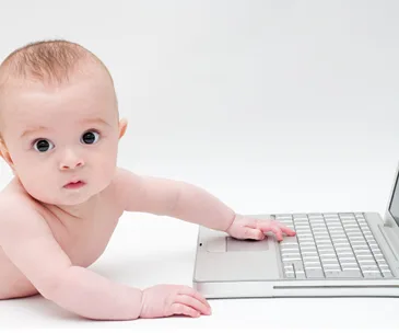 A baby with a curious expression reaches towards a laptop keyboard, exploring technology.