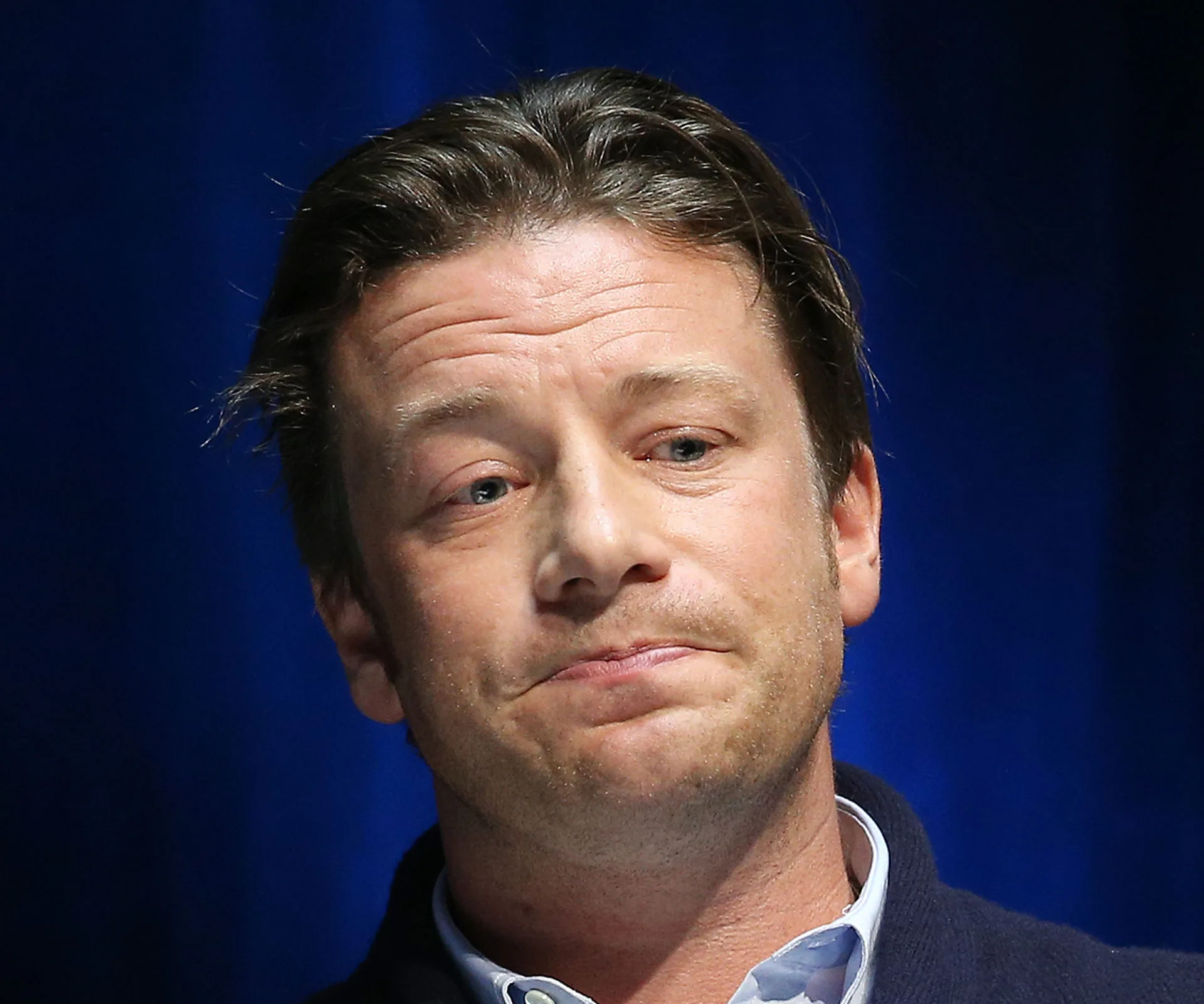 Chef speaking earnestly on stage, wearing a dark sweater, against a blue background.