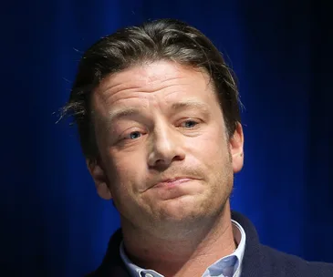 Chef speaking earnestly on stage, wearing a dark sweater, against a blue background.