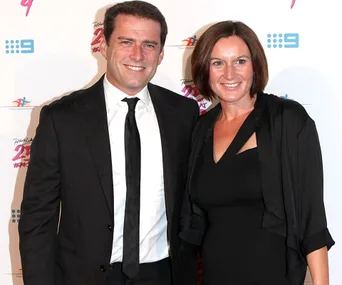 A man and woman smiling at a formal event, standing in front of a backdrop with logos and text.