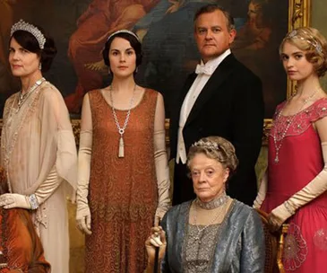 Cast of "Downton Abbey" in period costumes, posing elegantly in front of a painting.