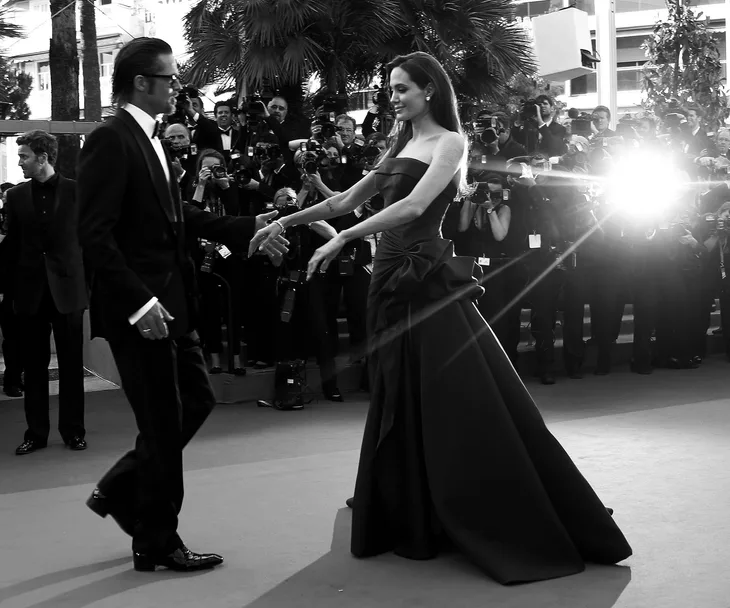 A couple in formal attire on a red carpet, surrounded by photographers and flashing cameras.