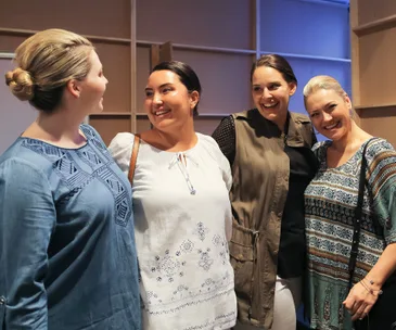 Four women standing together, smiling and conversing, dressed in casual attire.