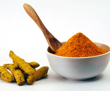 Bowl of turmeric powder with wooden spoon and whole turmeric roots on a white background.
