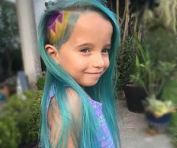 Girl with long turquoise hair and a side undercut featuring a colorful unicorn design, smiling outdoors.