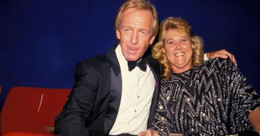 A man in a tuxedo and a woman in a sparkly dress sit together, smiling at the camera.