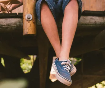 Child's legs dangling over wooden edge, wearing blue sneakers and denim shorts.